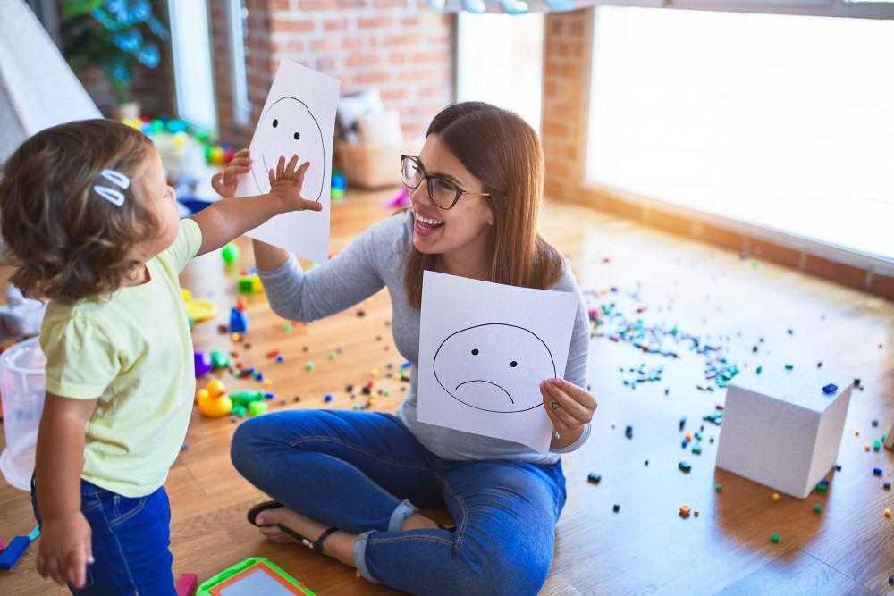 a teacher holds up two drawings of a smile and a sad face in front of a child, who is grabbing the happy face paper