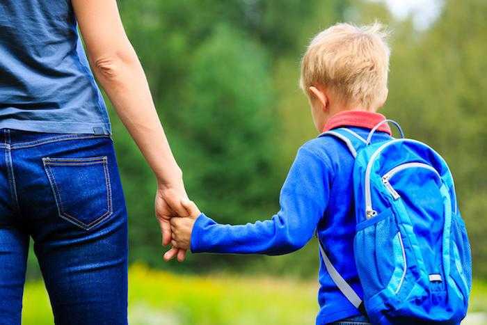 a young boy holds the hand of an adult with his backpack on