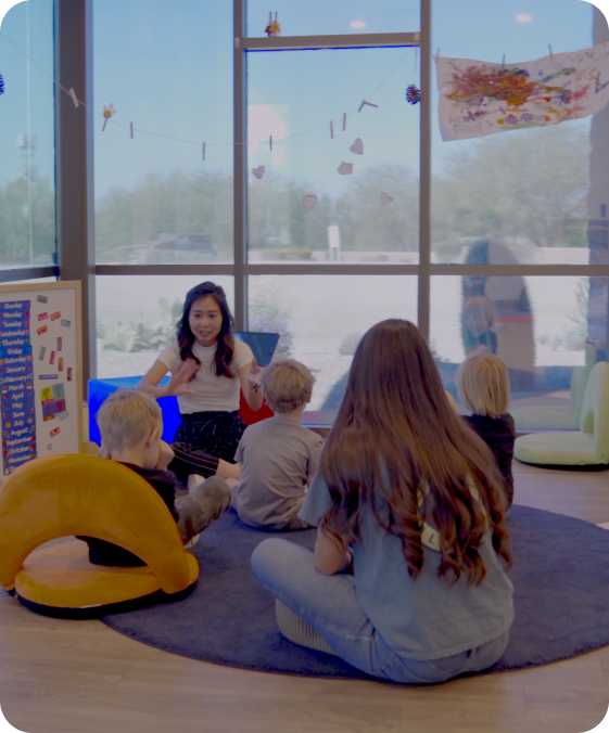 a teacher reads to a group of children sitting on the floor while two teachers supervise from behind