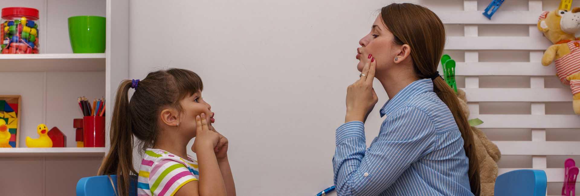 a woman and a child make a face to eachother while pressing their fingers into their cheeks, indicative of speech therapy