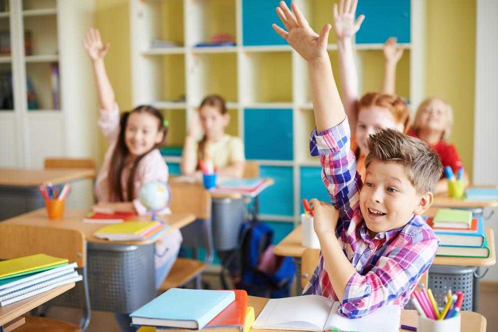 children raise their hands in a classroom setting at their desks