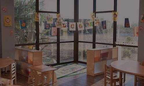 a room with bright windows and children's art on the window, surrounded by kids tables and chairs