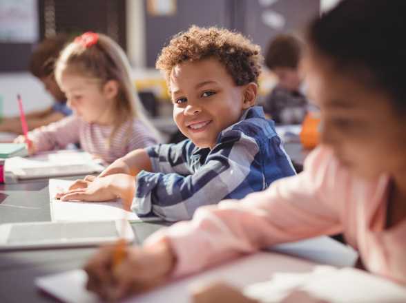 a little boy smiles at the camera, surrounded by other children who are reading and writing